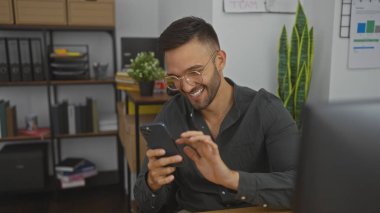 Yakışıklı, sakallı, genç, İspanyol bir adam ofisin içinde telefonuna bakarken gülümsüyor ve modern bir iş ortamı yaratıyor..