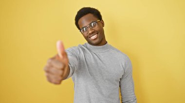Gözlüklü gülümseyen genç adam, parlak sarı bir arka plana başparmak sallıyor..