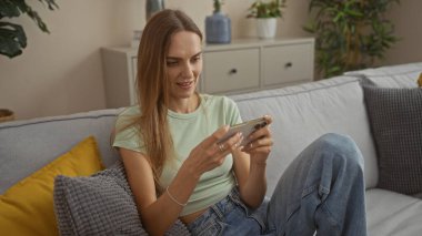 Güzel sarışın bir kadın oturma odasında dinleniyor, rahat gri bir kanepede otururken akıllı telefonuna bakıyor..