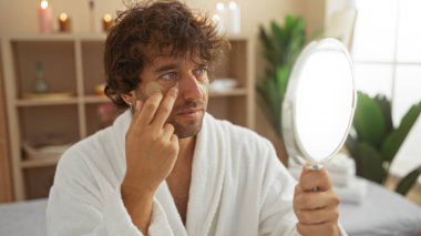 Beyaz bir bornoz giyen ve rahatlama salonunda ayna tutarken maske takan genç bir adam..