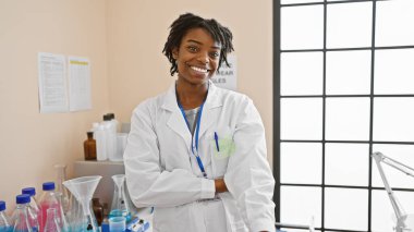 Bir bilim adamı gibi giyinmiş, rastalı saçlı, gülümseyen genç siyah kadın, laboratuvar ortamında güvenle duruyor..