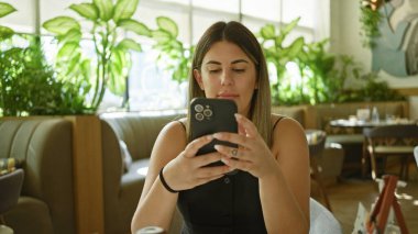 Çevresi yeşillik ve doğal ışıkla çevrili canlı bir kafede otururken akıllı telefon kullanan genç İspanyol kadın..