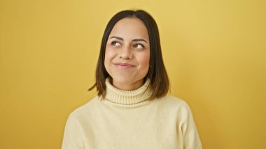 Boğazlı kazaklı, gülümseyen, genç bir İspanyol kadın canlı, sarı bir arka plana zarif bir şekilde poz veriyor..