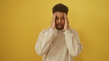 Sarı arka planı olan kapüşonlu stresli Afrikalı Amerikalı bir adam hayal kırıklığı ya da baş ağrısı içinde şakaklarına bastırıyor..