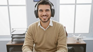 Modern ofis ortamında kulaklıklı, gülümseyen genç İspanyol adam, gündelik profesyonelliği yansıtıyor..