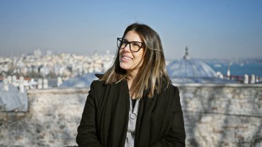 Güneşli bir günde İstanbul şehrine tepeden bakan siyah ceketli, gülümseyen kadın..