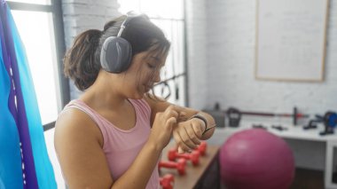 Esmer saçlı ve kulaklık takan Latin kökenli genç bir kadın spor salonunda spor salonunda spor aletleri ve arka planda spor topları gibi spor aletlerini kontrol ediyor..