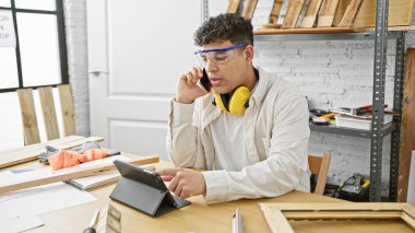 Güvenlik gözlüklü genç bir adam, ahşap ve aletlerle çevrili bir marangoz atölyesinde telefonla konuşuyor..