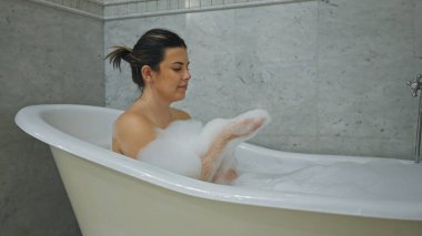 Rahatlamış genç bir kadın, beyaz bir küvette köpüklü bir banyonun keyfini çıkarıyor ve modern banyo düzenine huzur katıyor..
