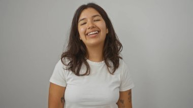 Genç, çekici, esmer saçlı ve dövmeli bir kadın izole edilmiş beyaz bir arka planda dikilirken neşeyle gülümsüyor..