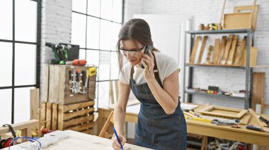 Parlak marangozluk atölyesinde ahşap işaretlerken güvenlik gözlüklü orta yaşlı bir kadın telefonla konuşuyor..