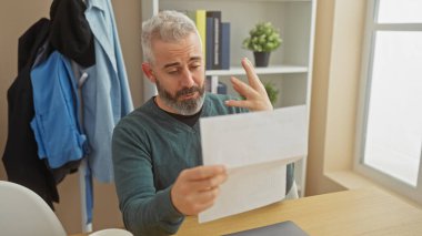 A perplexed man examines a document in a well-lit home office, reflecting a moment of everyday indoor life. clipart