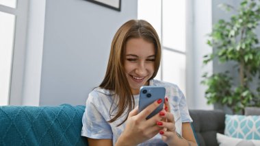 Gülümseyen beyaz bir kadın kanepede uzanıyor, rahat bir oturma odasında akıllı telefonunun keyfini çıkarıyor..