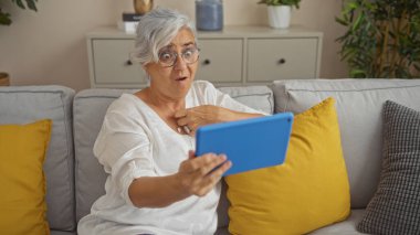 Oturma odasındaki kanepede oturan gri saçlı ve gözlüklü yaşlı kadın elinde mavi bir tablet tutarken şaşkın şaşkın bakıyor.