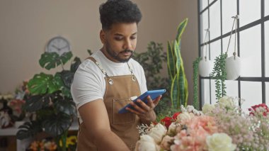 Afro-Amerikalı bir adam çiçekçi dükkanında tablet kullanıyor. Arka planda çeşitli bitkiler ve çiçekler var..