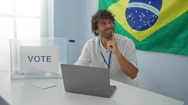 stock image Handsome young man sitting in an electoral college room in brazil with a ballot box and laptop, thinking while looking sideways