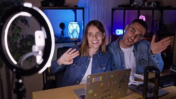 stock image A man and woman engage as content creators in a well-equipped, modern home office with a ring light, waving at their online audience.