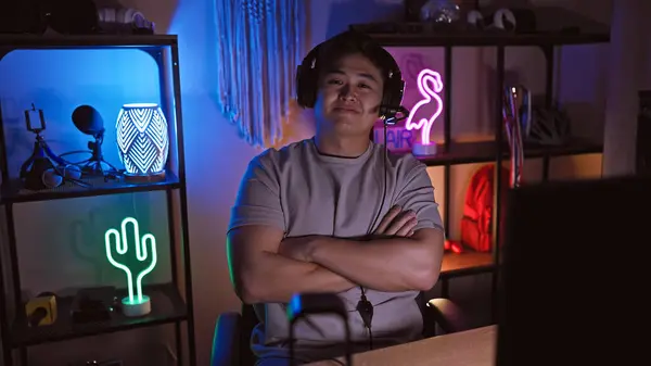 stock image A young asian man with crossed arms smiling indoors at night, surrounded by gaming room decor.