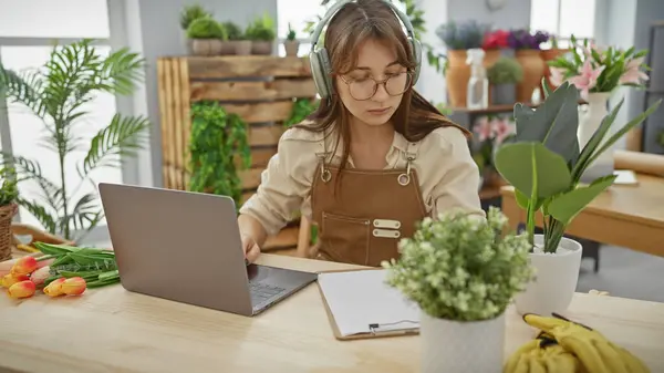 stock image A young woman florist with headphones uses a laptop in a plant-filled flower shop interior.