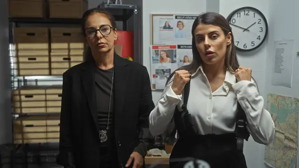 Two professional women detectives in a police station room, focused and prepared with badges, surrounded by investigation evidence.