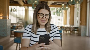 Modern bir kafenin içinde akıllı telefon kullanan gülümseyen genç bir kadın.