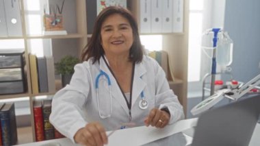 Kısa saçlı, beyaz önlüklü ve steteskoplu gülümseyen kadın doktor, tıbbi dosyalar ve ekipmanlarla bir klinikte oturuyor, dizüstü bilgisayar kullanırken gözlüğü ayarlıyor..