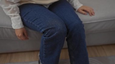 Orta yaşlı bir kadın evde oturuyor, elleri kotla kaplı dizlerinin üstünde ev ortamında görünüyor..