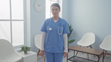 Steteskoplu mavi önlüklü genç bir kadın modern klinikte güvenle ayakta duruyor.