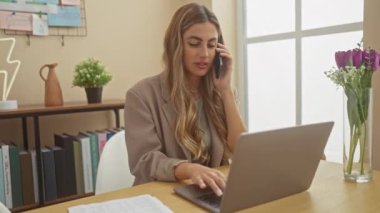 Evde çalışan odaklanmış sarışın bir kadın parlak bir oturma odasında dizüstü bilgisayar kullanırken telefonla konuşuyor..