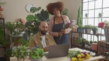 Çiçekçi bir kadın, çiçekçide elinde dizüstü bilgisayarla gülümseyen bir adama çiçek sunar..