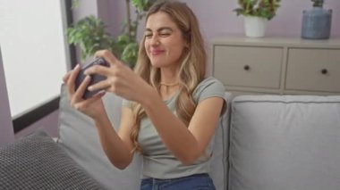 Gülümseyen beyaz bir kadın evde akıllı telefonuyla oyun oynuyor. Ev ortamında neşe ve heyecan ifade ediyor..