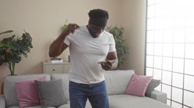 Genç, yakışıklı, sakallı Afrikalı Amerikalı bir adam oturma odasında neşeli bir şekilde dans ediyor. Elinde akıllı bir telefon var. Üzerinde gri bir kanepe ve pembe yastıklar var..