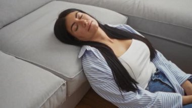 Uzun esmer saçlı İspanyol bir kadın evinin oturma odasındaki gri bir kanepede uzanır. Üzerinde çizgili bir gömlek ve kot pantolon vardır..