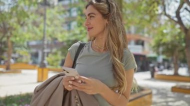 Sarı saçlı, gülümseyen genç bir kadın güneşli bir şehir parkında akıllı telefon kullanıyor. Bankları ve yeşil ağaçları var.