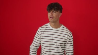 İspanyol asıllı genç adam, çizgili tişört giyiyor, ayakta duruyor ve uzaklara bakıyor, mutlu bir gülümsemeyle, el üstünde, izole edilmiş kırmızı bir arka planı arıyor.