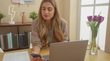 Modern bir ev ofisinde çiçek ve kitaplarla bilgisayar ve akıllı telefon kullanan odaklanmış beyaz bir kadın..