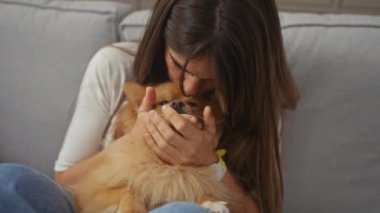 Genç bir kadın Pomeranian köpeğini nazikçe kucaklar, evdeki samimi ve sevgi dolu bir anı yansıtır..