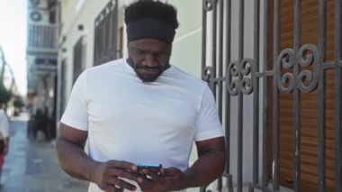 Yakışıklı, Afro-Amerikan bir adam şehir sokağında duruyor, telefonuna bakıyor, gün ışığında neşeli bir ifadeyle..