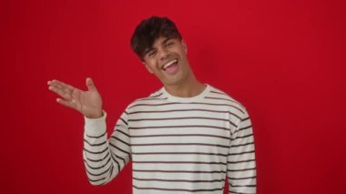 İspanyol asıllı genç adam, çizgili tişört giyiyor. Kırmızı arka planda duruyor. Dostane bir gülümseme ve mutlu bir jestle merhaba diyor.