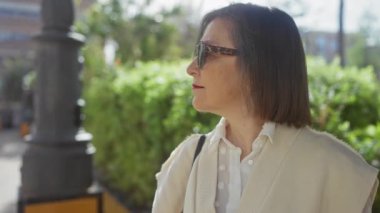 Güneş gözlüklü, düşünceli, olgun bir kadın arka planda yemyeşil bir şehir parkında duruyor..