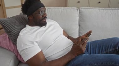 Yakışıklı Afro-Amerikalı sakallı bir adam evdeki koltukta dinleniyor, oturma odasında akıllı telefon kullanıyor, beyaz tişört ve kot pantolon giyiyor..
