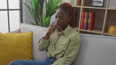 Afro-Amerikan bir kadın kendini kötü hissediyor ve modern bir oturma odasındaki koltukta öksürüyor.