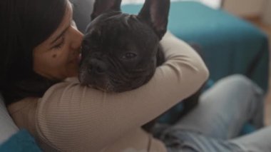 Genç bir İspanyol kadın, uykulu Fransız bulldog 'unu sıcak aydınlatılmış bir oturma odasında sıcak bir kanepede kucaklıyor. Sevgi ve dostluk dolu bir anı yakalıyor..