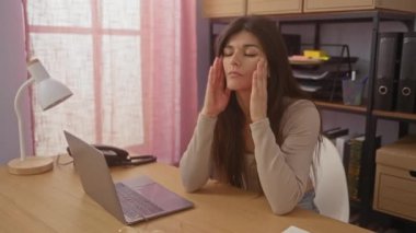 Güzel, esmer, İspanyol bir kadın ev ofis ortamında dizüstü bilgisayarında stresli bir şekilde beliriyor. Kapalı çalışma alanındaki işle ilgili baskıyı yakalıyor..