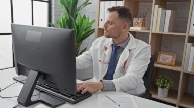 Odaklanmış genç bir adam, muhtemelen bir sağlık uzmanı, modern bir klinikte bilgisayar ve telefonla uğraşıyor..