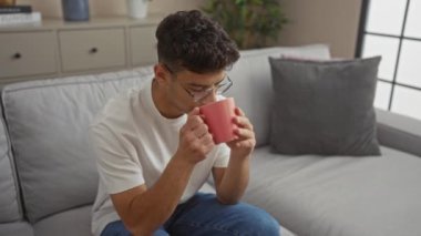 Genç İspanyol bir adam, oturma odasındaki rahat bir kanepede doğal bir dekor ve doğal ışıkla otururken pembe bardaktan sıcak bir içkinin tadını çıkarıyor..