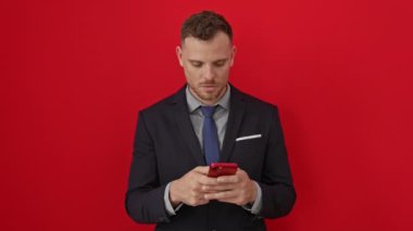 Şok olmuş ve korkmuş İspanyol kıyafetli genç adam kırmızı arka planda şaşırmış bir ifadeyle akıllı telefon kullanıyor.