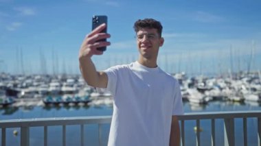 Beyaz tişörtlü genç İspanyol adam gülümsüyor ve güneşli bir günde arka planda çok sayıda tekne olan bir limanda akıllı telefonuyla selfie çekiyor..