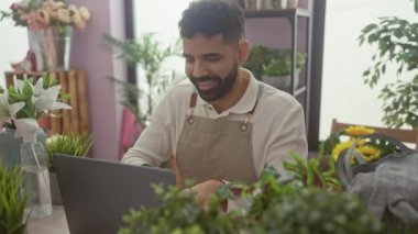 Sakallı yakışıklı, genç bir İspanyol, çiçek ve buketlerle çevrili bir çiçekçide dizüstü bilgisayarla çalışıyor..