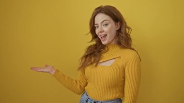 Süveter giyen genç beyaz kadın kameraya hayret içinde bakıp gülümserken eliyle ve parmağıyla işaret ediyor. izole edilmiş sarı arkaplan